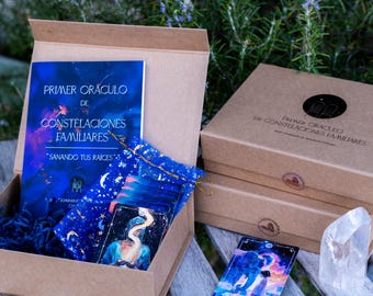 Puede incluir: Una caja marrón con una baraja de cartas azul y dorada, un libro titulado "Primer Oráculo de Constelaciones Familiares" y un cristal blanco. La caja está abierta y las cartas son visibles. El libro se titula "Primer Oráculo de Constelaciones Familiares" y tiene el texto "Sanando tus raíces" en la portada.