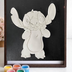 May include: A white, 3D-printed Stitch character art piece on a black background, framed in dark brown. The image includes small paint pots in various colors and a paintbrush, suggesting a DIY painting project. The artwork is a fun, creative item.