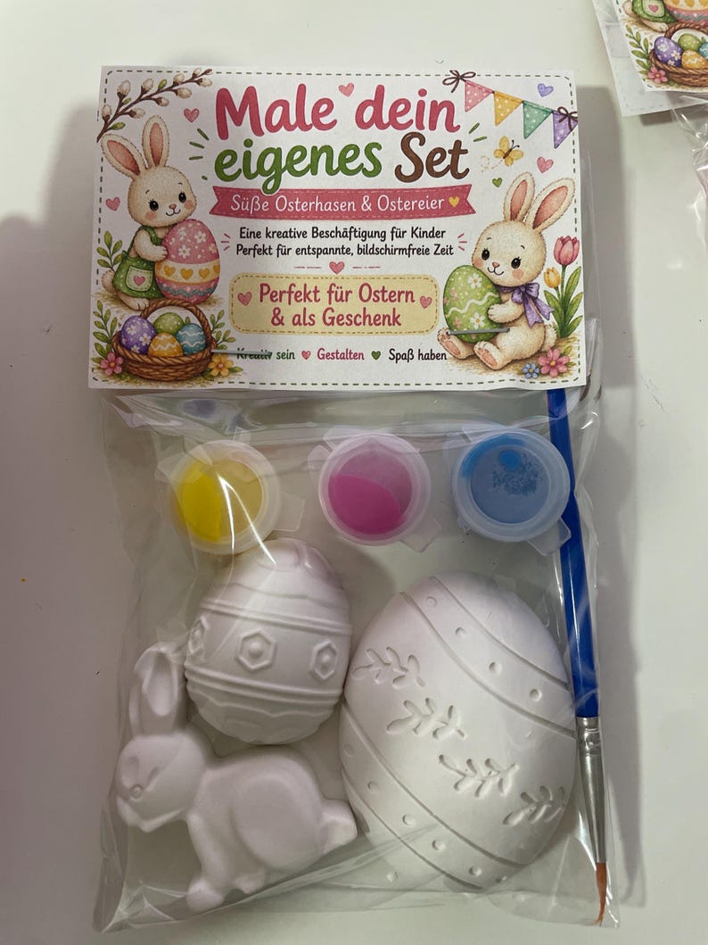 Puede incluir: Un juego de Pascua para pintar en una bolsa de pl&aacute;stico transparente. El juego incluye una figura de conejito, dos figuras en forma de huevo, botes de pintura y un pincel. El embalaje presenta ilustraciones de conejitos y huevos, con texto en alem&aacute;n.
