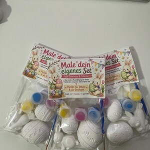Puede incluir: Tres juegos de pintura con tem&aacute;tica de Pascua. Cada juego incluye huevos y conejos de Pascua de cer&aacute;mica blanca, botes de pintura de varios colores y pinceles. El texto "Male dein eigenes Set" es visible.