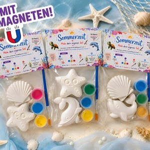 Op de afbeelding: Drie doe-het-zelf schilder kits met zeedieren. Elke kit bevat een witte keramische zeester, schelp en dolfijn, samen met verfpotten en een penseel. De verpakking bevat de tekst "Sommerzeit Male dein eigenes Set."