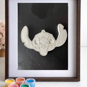 May include: A framed, white, 3D Stitch character art piece with a dark brown frame. The artwork is displayed on a black background. Paint pots in yellow, orange, pink, green, and blue, along with a paintbrush, are in the foreground.