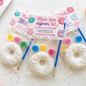 Op de afbeelding: Drie doe-het-zelf donut kits met witte, ongeverfde donuts, verfpotjes en penselen. Elke kit bevat een donut, verfpotjes in verschillende kleuren en een blauwe penseel. De kits zijn verpakt in doorzichtige plastic zakken.