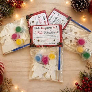 Könnte beinhalten: Weihnachtliche Bastelsets mit Farbe und Pinseln. Jedes Set enthält weiße Gipsornamente in verschiedenen Formen, darunter ein Weihnachtsbaum, ein Schneeflocke und eine Weihnachtsmütze. Die Sets enthalten rote, rosa, gelbe und grüne Farbe.