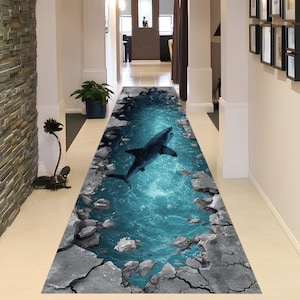 May include: A long, rectangular rug with a 3D shark design. The rug features a vibrant blue ocean scene with a shark swimming through it, surrounded by a cracked, rocky border. The rug is designed to create an illusion of depth.