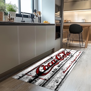May include: A white and gray kitchen runner rug with a red and white Coca-Cola logo. The rug has a distressed woodgrain pattern and the text "Drink Coca-Cola in Bottles 6.5 fl. oz. American's Original Since 1886".