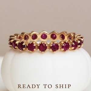 Pode incluir: Um anel de banda de ouro com uma fileira de pedras de rubi vermelho. O anel está sobre um pedestal branco com o texto "READY TO SHIP".