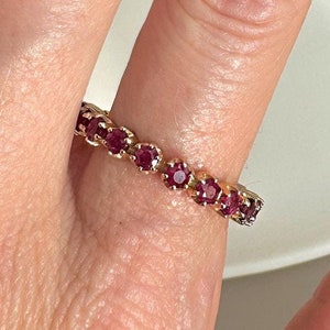 Pode incluir: Um anel de banda de ouro com uma fileira de pequenas pedras preciosas vermelhas.