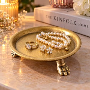 May include: A round gold-toned metal jewelry tray with three claw-shaped feet. The tray holds a pearl bracelet, a gold ring with a central clear stone, and a pair of gold flower-shaped stud earrings with clear stones.