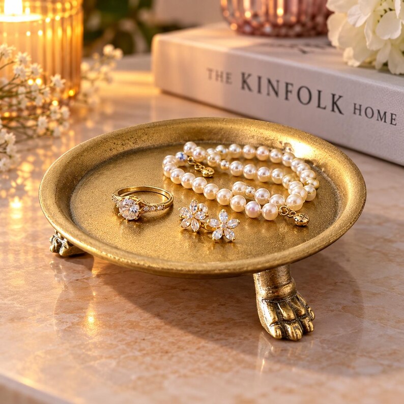 May include: A round gold-toned metal jewelry tray with three claw-shaped feet. The tray holds a pearl bracelet, a gold ring with a central clear stone, and a pair of gold flower-shaped stud earrings with clear stones.