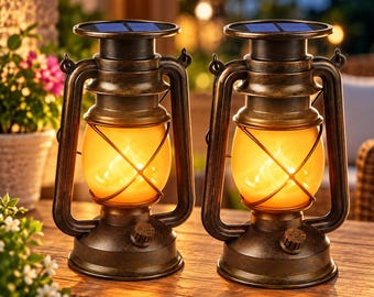 May include: Two antique-style bronze-colored metal lanterns featuring solar panels on top and amber-colored glass globes that emit a warm, flickering light. Each lantern has a wire cage protecting the glass and a small knob on the base.