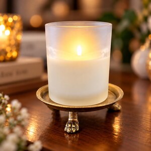 May include: A frosted glass candle holder containing a lit candle with a warm glow, resting on a circular, antique gold-toned metal stand featuring three detailed claw-shaped feet that provide a stable base for the cylindrical vessel.