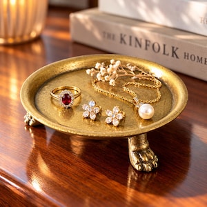 May include: A round, gold-toned metal pedestal tray with three claw-shaped feet. Inside the tray sits a gold ring with a central oval red gemstone surrounded by small clear stones, two flower-shaped stud earrings, and a gold chain necklace.
