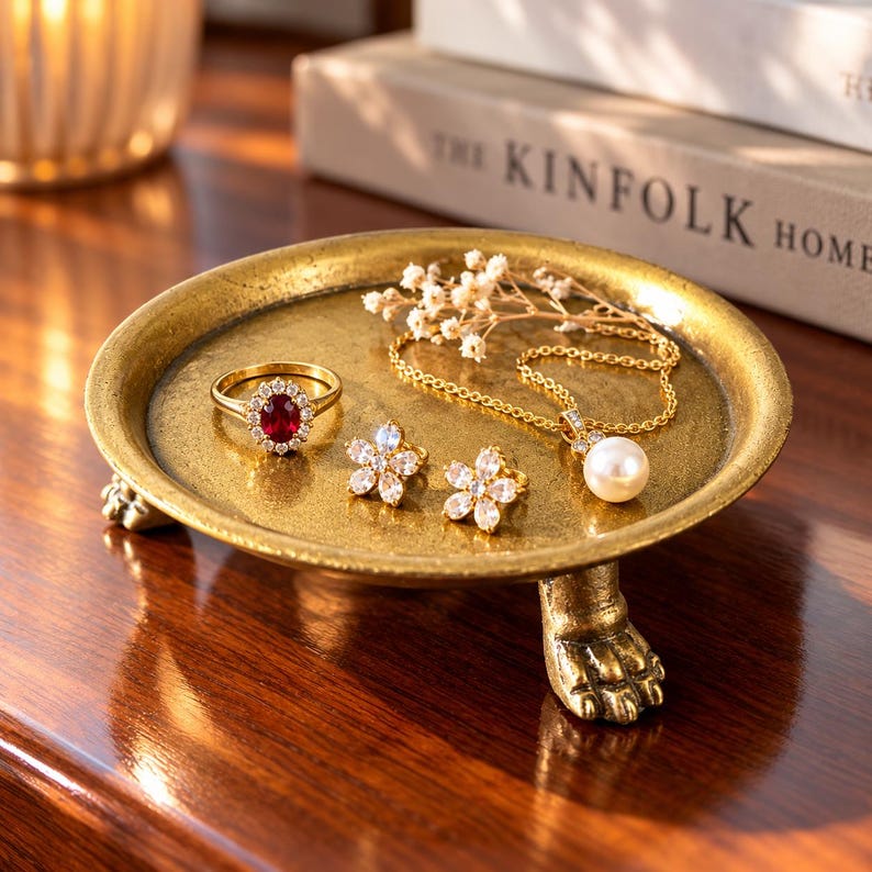 May include: A round, gold-toned metal pedestal tray with three claw-shaped feet. Inside the tray sits a gold ring with a central oval red gemstone surrounded by small clear stones, two flower-shaped stud earrings, and a gold chain necklace.