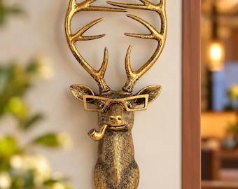 May include: A wall-mounted deer head sculpture finished in a metallic gold tone with dark antiqued highlights. The deer features large antlers, wears rectangular-framed glasses, and has a pipe protruding from the side of its mouth.