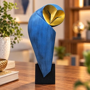 May include: A stylized owl sculpture featuring a smooth, deep blue body with a textured finish and a contrasting metallic gold face. The figure is mounted on a solid, square black base, creating a sleek and modern decorative accent piece.