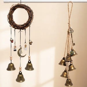 May include: A hanging wall decoration featuring a circular twig wreath with dangling black cords, brass bells, small glass vials, and a crescent moon charm, alongside a separate vertical cluster of brass bells suspended by jute twine with a key.