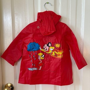 May include: A bright red raincoat featuring a hood and a graphic print on the back. The illustration depicts a cartoon mouse character flying toward a blue rain cloud above a small figure with yellow hair, accompanied by a smiling sun icon.