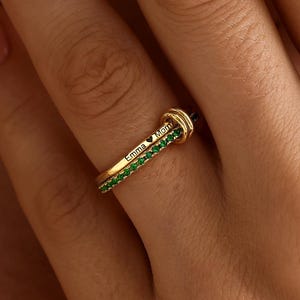 May include: A gold ring featuring two thin bands. One band is engraved with the text Emma and a heart symbol, while the other band is set with a row of small, vibrant green stones. The two bands are linked together by a small gold loop.