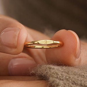 Könnte beinhalten: Ein goldener Ring mit einem kleinen, rechteckigen Design auf der Oberseite, mit dem Buchstaben "R". Der Ring wird zwischen den Fingern gehalten und zeigt seine polierte Oberfläche. Der Hintergrund ist eine weiche, neutrale Farbe.