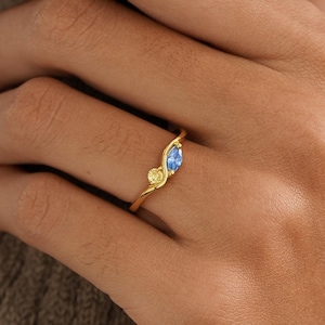 Peut inclure: Une bague en or ornée d'une petite pierre jaune ronde et d'une pierre bleue ovale légèrement plus grande, fixées sur un anneau fin et ondulé. Le design intègre deux pierres distinctes disposées dans un style croisé, avec une finition dorée.