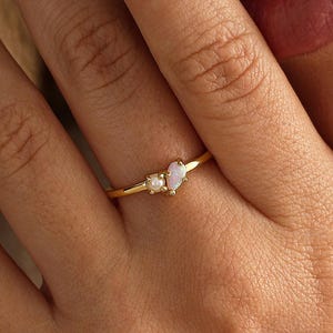 Puede incluir: Un anillo de oro con una pequeña piedra redonda y una piedra iridiscente en forma de marquesa sobre una banda fina. El anillo está colocado en un dedo, mostrando el delicado trabajo del metal y las dos formas distintas de las piedras.