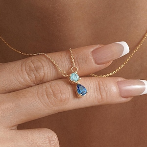 Puede incluir: Un delicado collar de oro con una pequeña gema redonda azul claro y un colgante en forma de lágrima azul oscuro. El collar se muestra sobre un fondo neutro, resaltando el diseño y el color de la joya.