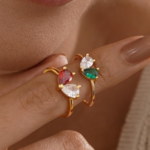 May include: Two gold rings, each with a cluster of pear-shaped gemstones. One ring features red and white stones, the other green and white. The rings are displayed on a finger, showcasing the jewelry's design and colour.