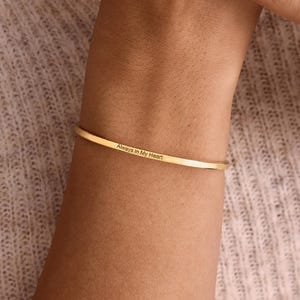 May include: A thin, gold-toned metal cuff bracelet with a smooth, polished finish. The exterior surface of the bracelet features the engraved text "Always in My Heart" in a simple, clean, dark-coloured sans-serif font centred on the band.
