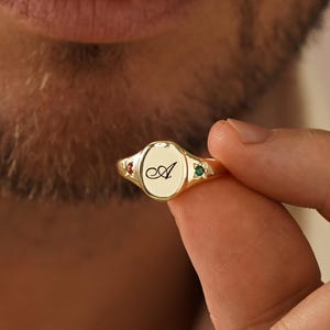 May include: A gold-toned signet ring featuring an oval face engraved with the cursive letter A. The ring band is accented on both sides by small, star-shaped settings containing a single red gemstone on the left and a green gemstone on the right.