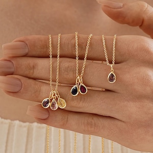 May include: Gold-toned necklaces with teardrop-shaped gemstones in various colors, including purple, pink, and yellow. The necklaces are displayed on a hand, showcasing the delicate chains and pendants. The gemstones are approximately 0.5 inches in length.