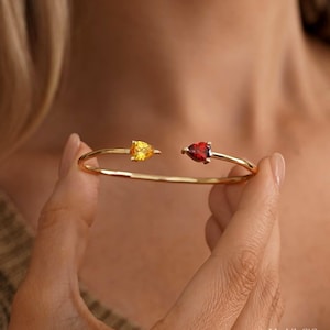 May include: A gold-toned open cuff bracelet featuring two pear-shaped gemstones at the ends. One gemstone is a vibrant yellow, while the other is a deep red. The thin, polished metal band creates a delicate, minimalist silhouette for the wrist.