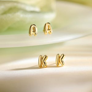 May include: A pair of gold-toned stud earrings shaped like the letter A and a pair of gold-toned stud earrings shaped like the letter K. Each earring features a smooth, rounded, puffed surface with a polished finish and a post-back closure.