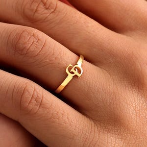 Könnte beinhalten: Ein goldfarbener Ring mit einem zentralen Monogramm-Design, bei dem die Buchstaben D und G ineinander verschlungen sind. Das Band ist dünn und poliert und weist eine metallische Oberfläche auf, die das Licht reflektiert.