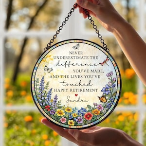 May include: A round, stained-glass retirement plaque with a black chain for hanging. The plaque features colorful flowers, butterflies, and the text "NEVER UNDERESTIMATE THE difference YOU'VE MADE, AND THE LIVES YOU'VE touched HAPPY RETIREMENT Sandra."