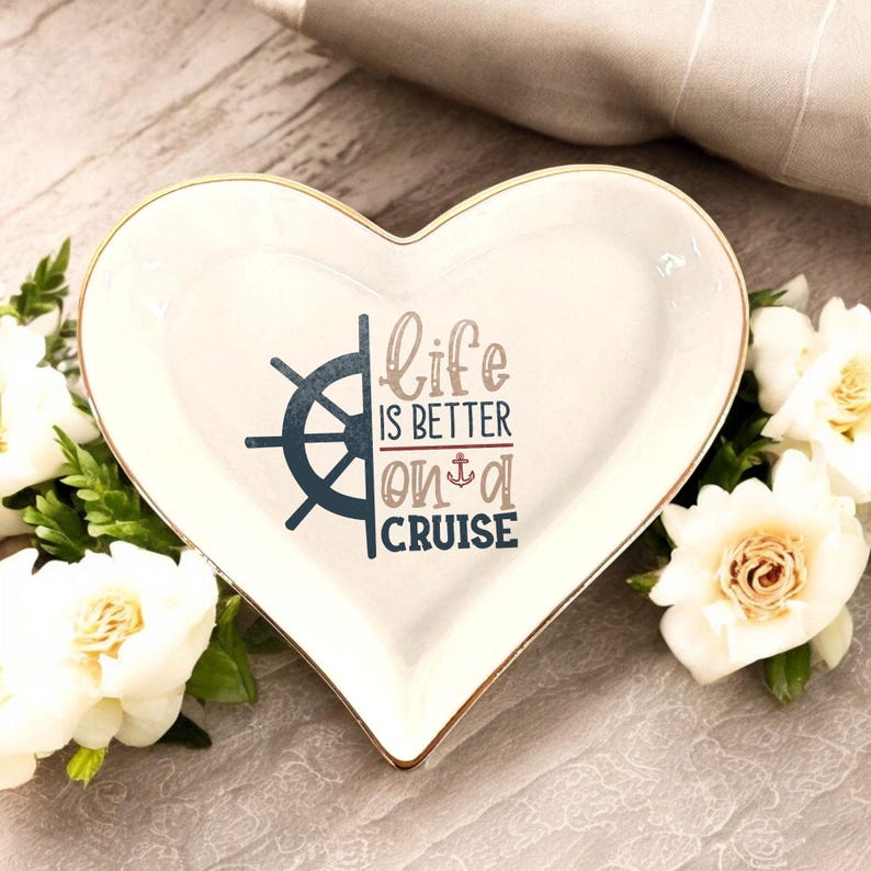 May include: White heart-shaped ceramic trinket dish with a gold rim. The dish features a blue and white nautical wheel and the text "life is better on a cruise".