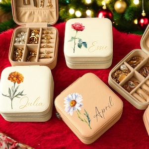 May include: Several open jewelry travel cases with gold jewelry and pearls. The cases are beige with floral designs and names like "Essie" and "April". The cases are on a red surface with Christmas decorations in the background.