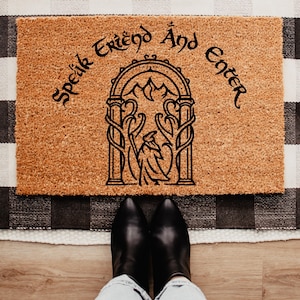 May include: A rectangular doormat with the text "Speak Friend And Enter" in black script. The mat features a black illustration of a figure in an archway with mountains in the background. The mat is made of a light brown material and is placed on a black and white checkered rug.