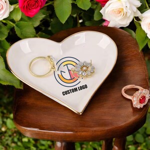 May include: White heart-shaped ceramic trinket dish with a gold rim. The dish has a custom logo printed on it and is sitting on a wooden surface. There are three pieces of jewelry on the dish: a gold ring, a gold hoop earring, and a silver flower-shaped earring.