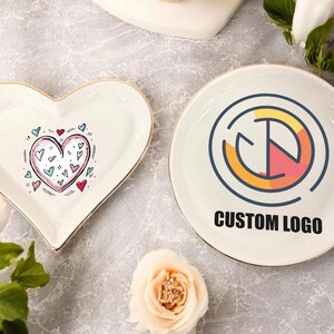 May include: Two white ceramic dishes, one shaped like a heart with a pink and blue heart design and the other round with a gold and red logo that says "CUSTOM LOGO".
