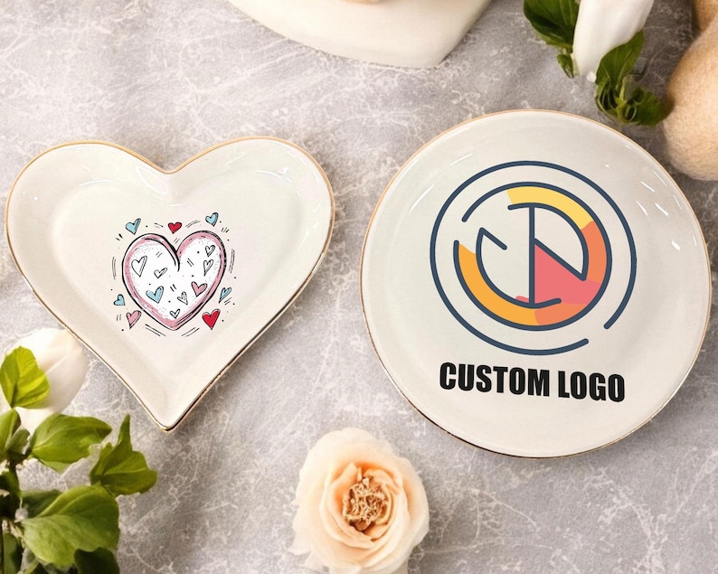 May include: Two white ceramic dishes, one shaped like a heart with a pink and blue heart design and the other round with a gold and red logo that says "CUSTOM LOGO".