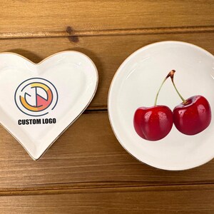 May include: Two white ceramic dishes, one heart-shaped and one round. The heart-shaped dish has a gold rim and a graphic design with the text "CUSTOM LOGO". The round dish has a gold rim and a design of two red cherries.