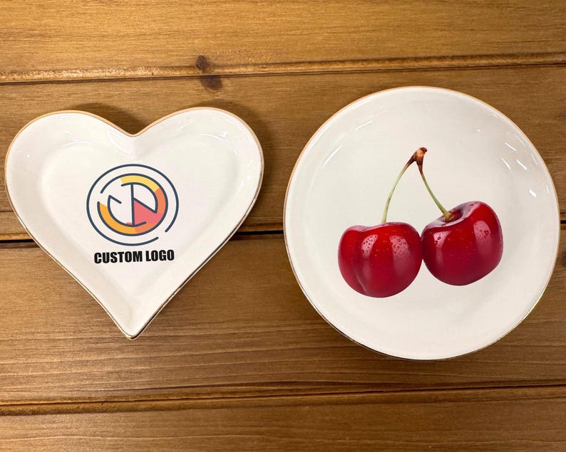 May include: Two white ceramic dishes, one heart-shaped and one round. The heart-shaped dish has a gold rim and a graphic design with the text "CUSTOM LOGO". The round dish has a gold rim and a design of two red cherries.