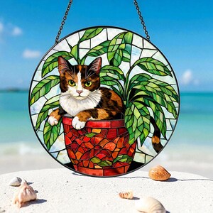 May include: Round stained glass window featuring a calico cat in a red mosaic pot with green leaves. The cat has brown, white, and orange fur and green eyes. The background is a blue sky and ocean.