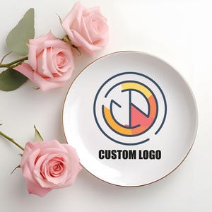 May include: White ceramic plate with a gold rim, featuring a colorful logo with the text "CUSTOM LOGO". The logo is a circle with a yellow and red design inside.