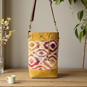 Puede incluir: Bolso tipo cubo con un estampado geométrico en tonos naranja, rosa y morado. El bolso tiene una base y parte superior de ante amarillo mostaza, con una correa de cuero marrón y cierre de cordón. El bolso mide unos 30 cm de alto.