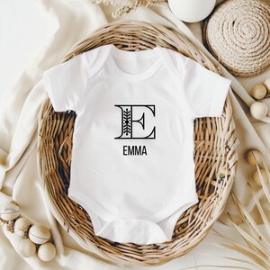 Könnte beinhalten: Weißer Baby-Strampler mit einer schwarzen Grafik des Buchstabens E mit einem floralen Design im Buchstaben und dem Namen "EMMA" darunter.