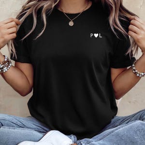 Könnte beinhalten: Schwarzes T-Shirt mit den Buchstaben "P" und "L", getrennt durch ein weißes Herz. Das Shirt hat kurze Ärmel und ist aus weichem Material. Es wird mit blauen Jeans und weißen Turnschuhen getragen.