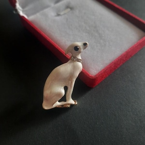 Può includere: Una spilla in smalto color crema a forma di levriero seduto. Il cane presenta un occhio nero e un collare decorato con piccoli strass trasparenti. La spilla è posizionata davanti a un portagioie rosso aperto con rivestimento interno bianco.