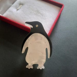 May include: A penguin-shaped brooch featuring a glossy black head and back with a pearlescent white belly. The design includes a small, clear circular rhinestone accent positioned on the head to represent an eye, crafted from smooth acrylic material.